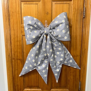 May include: A large, fabric bow featuring a gray background with a repeating pattern of small white daisies with yellow centers. The bow has two wide top loops and two long, pointed tails hanging downward, constructed from a quilted material.
