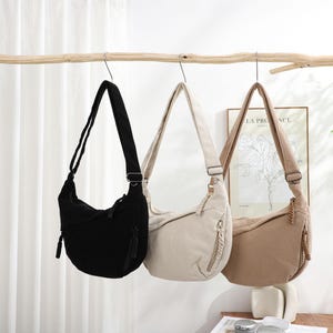 May include: Three corduroy shoulder bags in black, cream, and tan hang from a wooden branch. Each bag has a long adjustable strap and a zippered front pocket. The bags are displayed against a neutral background.