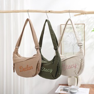 May include: Three corduroy shoulder bags in beige, olive green, and cream hang from a branch. Each bag has a long strap, a zipper, and an embroidered name: "Declan," "Stacy," and "Stacy." The bags are a casual style.