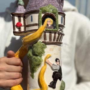 以下が含まれることがあります： 紫色の屋根と黄色の編み込みの取っ手を持つ塔の形をしたセラミックマグカップ。塔には窓があり、長い金髪の女の子が外を見ている。男性が編み込みを登っている。