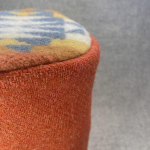 Puede incluir: Primer plano de un otomán cilíndrico naranja. La parte superior presenta un diseño en azul, amarillo y blanco. El otomán tiene una apariencia texturizada y tejida, y los bordes están cuidadosamente acabados.