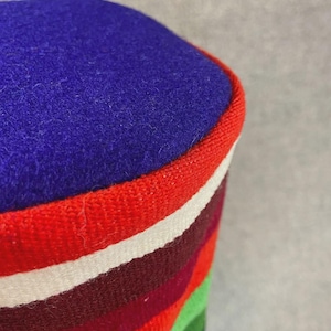Op de afbeelding: Een cilindrisch textielitem met een levendig ontwerp. De bovenkant is diepblauw, afgezet met een rode rand. De body heeft horizontale strepen in rood, wit, bordeaux, groen en andere kleuren. Waarschijnlijk een decoratief kussen of poef.