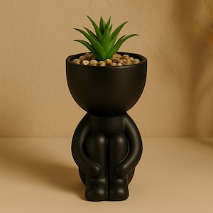 Puede incluir: Un macetero de cerámica negro con forma de persona sentada con las piernas cruzadas, sosteniendo una pequeña planta suculenta verde.