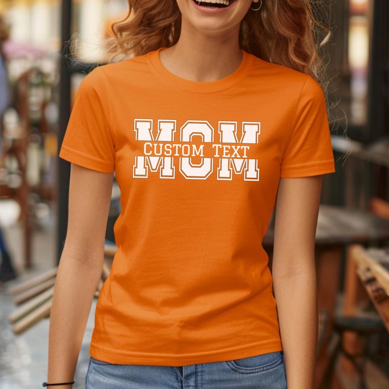 Może przedstawiać: Pomarańczowa koszulka z napisem "MOM" w białych blokowych literach, z napisem "CUSTOM TEXT" powyżej i "MOM" poniżej. Koszulka jest kr&oacute;tka i ma okrągły dekolt.
