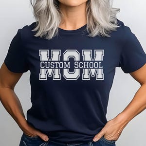 Puede incluir: Camiseta azul marino con el texto "MOM CUSTOM SCHOOL MOM" en blanco, con una fuente de estilo universitario. El texto está centrado en la parte delantera de la camiseta. La camiseta tiene cuello redondo y mangas cortas.