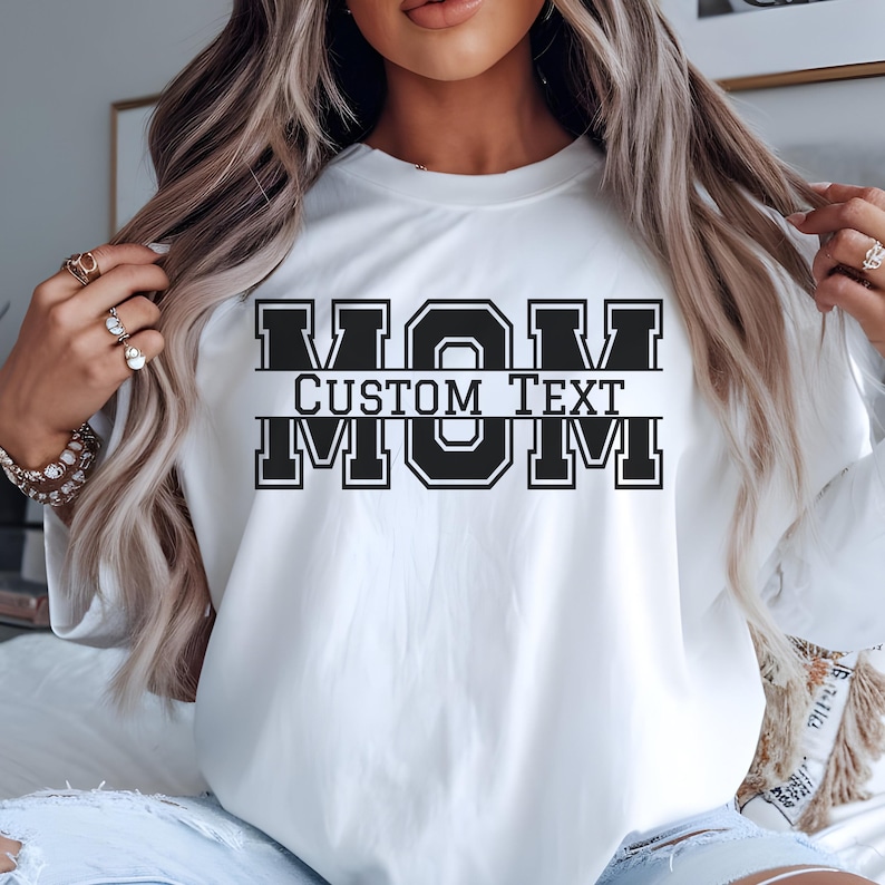 Może przedstawiać: Biała bluza z napisem "MOM" pogrubioną, czarną czcionką w stylu uniwersyteckim. Słowa "CUSTOM TEXT" są wyśrodkowane pod słowem "MOM". Bluza ma okrągły dekolt i długie rękawy.
