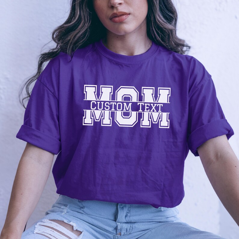 Może przedstawiać: Fioletowa koszulka z napisem "MOM" w czcionce w stylu uniwersyteckim. Tekst "CUSTOM TEXT" znajduje się pomiędzy dwoma słowami "MOM". Koszulka wykonana jest z miękkiego materiału i nadaje się do noszenia na co dzień.