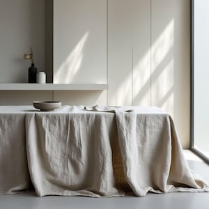 Op de afbeelding: Een beige linnen tafelkleed ligt over een rechthoekige tafel en heeft een gestructureerd stoffen oppervlak. Een kleine, ondiepe kom met een matte afwerking staat op de tafel. Op de achtergrond staan een zwarte fles en een witte houder op een plank.