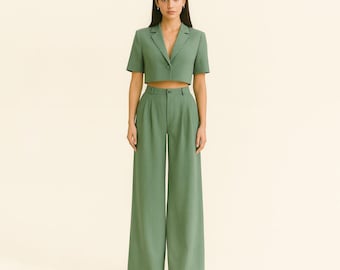 May include: A two-piece sage green suit. The cropped blazer has short sleeves and a single button closure. The high-waisted, wide-leg pants have a pleated front. The outfit is made of a solid-colored fabric.