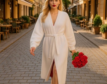 Pode incluir: Cardigã longo branco com cinto, decote em V e mangas compridas. O cardigã é usado sobre um vestido vermelho e combinado com sapatos brancos. Um buquê de rosas vermelhas é segurado na mão.
