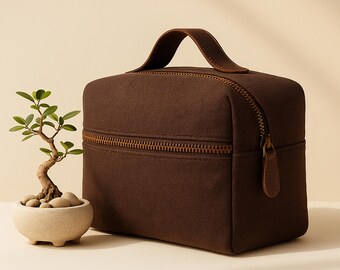 Op de afbeelding: Een bruine rechthoekige reistas met een handvat aan de bovenkant en een ritssluiting rondom. De tas heeft een leren handvat en ritstrekker. Een kleine bonsai in een crèmekleurige pot staat links.