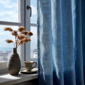May include: A window with a blue linen curtain, a ceramic vase with dried flowers, and a cup and saucer. The curtain is a solid light blue color. The vase is a dark gray with horizontal lines. The cup and saucer are light brown.