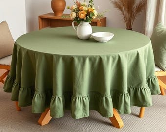 May include: A round, sage green tablecloth with a ruffled edge. A white vase holds a bouquet of flowers on the table. A white bowl sits next to the vase. The table is made of wood.