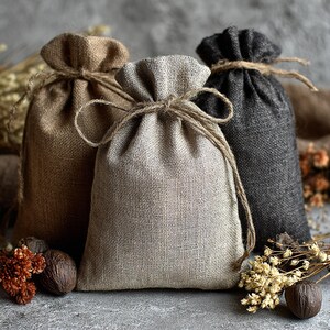 May include: Three burlap drawstring bags in shades of brown and gray. Each bag is tied with twine at the top. The bags are arranged on a gray surface with dried flowers and nuts.
