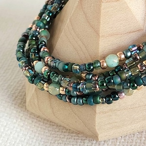 May include: A multi-strand bracelet made of small green, blue, and copper beads. The bracelet is wrapped around a wooden display stand.