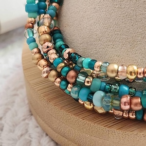 May include: Close-up of several beaded bracelets in shades of teal, turquoise, gold, and copper. The bracelets are wrapped around a wooden form, showcasing the variety of bead sizes and textures. The beads are strung together, creating a layered effect.