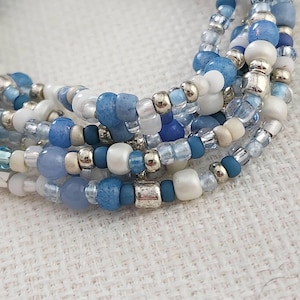 May include: A multi-strand bracelet composed of various beads in shades of blue, white, and silver. The design features a mix of matte, glossy, translucent, and metallic beads arranged in a layered pattern, creating a textured appearance.