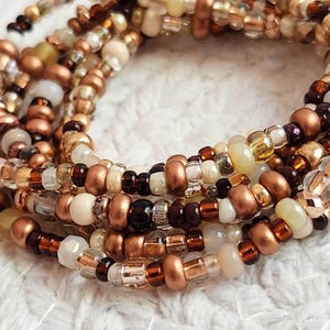 May include: Close-up of several strands of beaded jewelry. The beads are a mix of sizes and shapes, in shades of copper, gold, brown, cream, and clear. The jewelry is arranged in a spiral pattern.