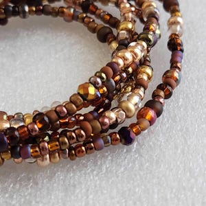 May include: Close-up of multiple strands of beaded bracelets. The beads are in shades of brown, gold, and purple. The beads vary in size and shape, with some faceted and others smooth. The bracelets are arranged in a layered style.