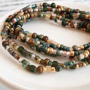 May include: A multi-strand beaded bracelet with green, gold, and brown beads. The bracelet is made of small, round beads and is wrapped around a white surface.