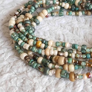 May include: A close-up of a multi-strand beaded necklace with a mix of cream, beige, gold, and teal beads.