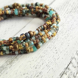 May include: A multi-strand beaded bracelet featuring a mix of small, colorful beads in shades of turquoise, brown, gold, and cream. The bracelet is coiled and rests on a white wooden surface, showcasing the intricate details of the jewelry.