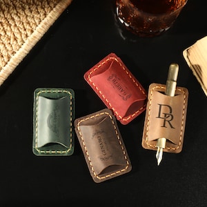 Puede incluir: Cuatro estuches de cuero para bolígrafos en verde, rojo, marrón y beige, con costuras en contraste. Un estuche contiene un bolígrafo. Los estuches tienen texto e iniciales en relieve. Los artículos están sobre una superficie negra.
