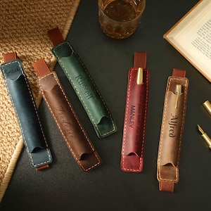 May include: A collection of personalized leather pen holders in various colors: navy, brown, green, burgundy, and tan. Each holder features a name engraved on the front. A pen is visible in two of the holders. The holders are on a dark surface with a book and a glass of liquid.