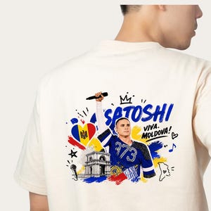 Könnte beinhalten: Ein cremefarbenes T-Shirt mit einem Grafikdruck auf der Rückseite, der den Künstler Satoshi mit einem Mikrofon zeigt. Das Design enthält die moldauische Flagge, den Triumphbogen, den Text "SATOSHI" und "VIVA. MOLDOVA!" sowie blaue und rote Farbspritzer.