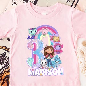 May include: A light pink toddler t-shirt featuring a colorful graphic with the number 3, a rainbow, and cartoon characters. The name "MADISON" is printed below the graphic in purple letters. The shirt is made of soft material.