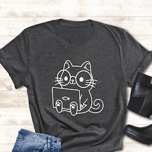 May include: Dark gray t-shirt with a white graphic of a cat wearing glasses and using a laptop. The cat graphic has a fish on the laptop screen. The image also includes blue jeans, sunglasses, and black ankle boots.