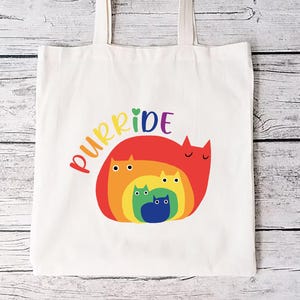 May include: A white canvas tote bag with the word "purride" in rainbow colors and a rainbow-colored cat design. The bag has two handles and is made of a sturdy fabric, perfect for carrying groceries or books.