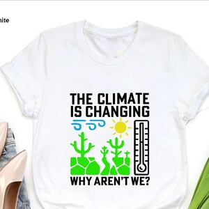 May include: White t-shirt with the text "THE CLIMATE IS CHANGING WHY AREN'T WE?" in black, with illustrations of a sun, thermometer, and cacti in green, blue, and yellow. The shirt is a crew neck.