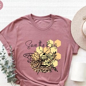 May include: Mauve t-shirt with a graphic of a bee with a leopard print body, floral accents, and the text "Save the Bees." The bee is set against a yellow honeycomb design. A white mug and a beige hat are visible.