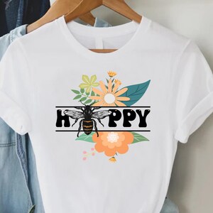 May include: White t-shirt with a graphic design featuring a bee and the word "HAPPY" in black letters. The design includes floral elements in shades of orange, green, and yellow. The shirt is hanging on a wooden hanger with other clothing items.