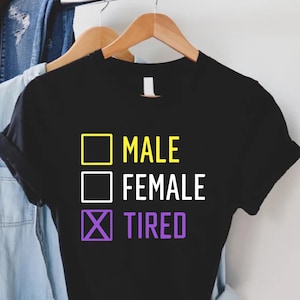 May include: Black t-shirt with a white graphic that says "Male", "Female", and "Tired" with a checkmark next to "Tired" in purple and yellow.