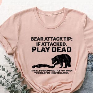 May include: A light pink t-shirt with the text "BEAR ATTACK TIP: IF ATTACKED, PLAY DEAD" in black. Below the text is a graphic of a bear and a person. The text "IT WILL BE GOOD PRACTICE FOR WHEN YOU DIE A FEW MINUTES LATER" is below the graphic.