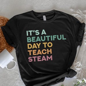 May include: Black t-shirt with the text "IT'S A BEAUTIFUL DAY TO TEACH STEAM" in teal, yellow, orange, and pink. The shirt is displayed on a white surface with a woven basket, a white mug, and flowers.