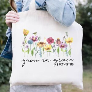 May include: Cream-colored tote bag featuring a watercolor floral design with yellow, pink, and blue flowers. The words "grow in grace" and "2 PETER 3:18" are written in black script. The bag is held by a person wearing a denim jacket.