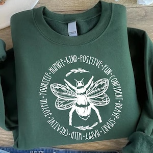 May include: A dark green sweatshirt with a white graphic of a bee inside a circle of words. The words include "Humble, Kind, Positive, Fun, Confident, Brave, Smart, Happy, Wild, Creative, Joyful, Yourself." The sweatshirt is folded.