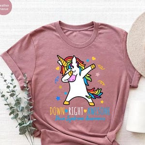 May include: A heather mauve t-shirt with a rainbow unicorn dabbing and the text "Down Right Awesome Down Syndrome Awareness".
