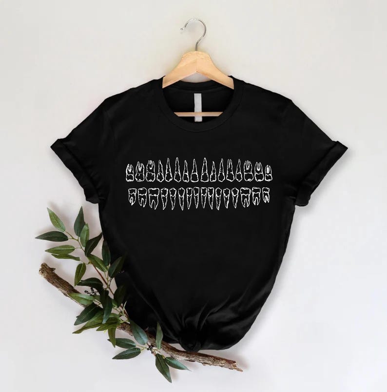 May include: Black t-shirt featuring a white graphic of a full set of human teeth. The teeth are arranged in a row, showing the different types of teeth. The shirt is hanging on a wooden hanger against a white background.