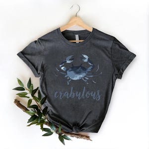 以下が含まれることがあります： 青いカニのグラフィックと「crabulous」の文字が書かれた濃い灰色のTシャツ。木製のハンガーにTシャツがかけられています。枝と緑の葉が左下に配置されています。