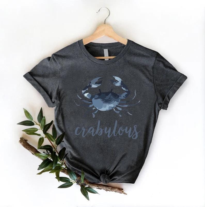 以下が含まれることがあります： 青いカニのグラフィックと「crabulous」の文字が書かれた濃い灰色のTシャツ。木製のハンガーにTシャツがかけられています。枝と緑の葉が左下に配置されています。