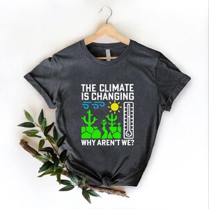 May include: Dark gray t-shirt with the text "THE CLIMATE IS CHANGING WHY AREN'T WE?" and a graphic of a sun, thermometer, and cacti. The shirt is hanging on a wooden hanger.