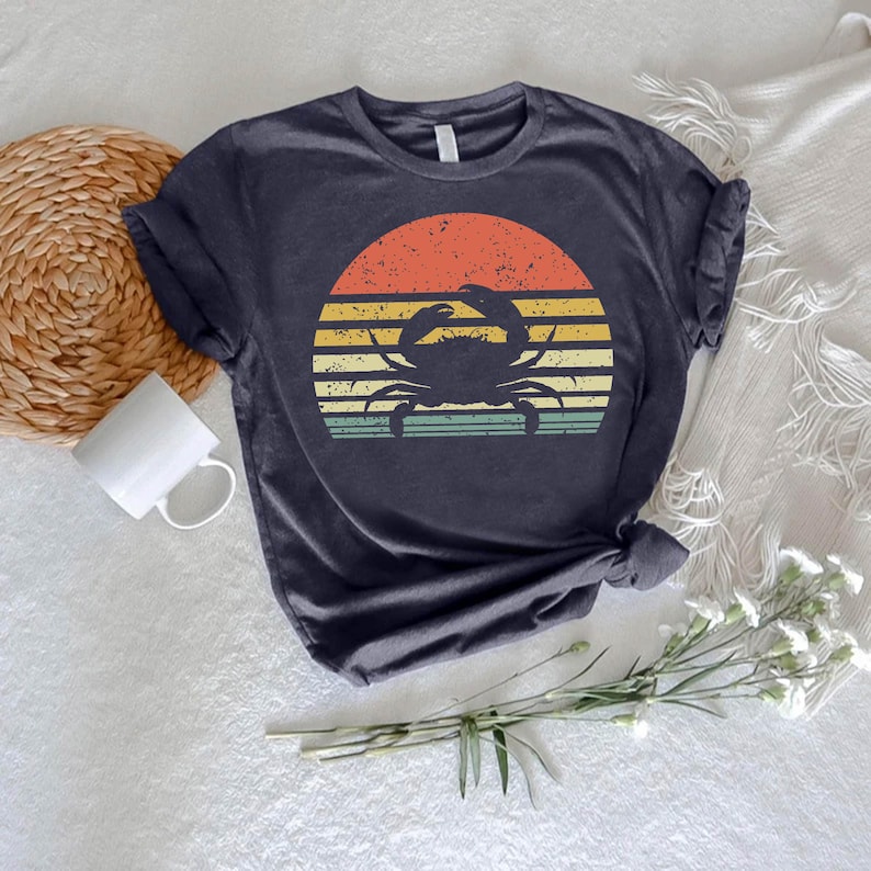 May include: Navy blue t-shirt featuring a crab silhouette against a retro sunset design. The sunset has horizontal stripes in orange, yellow, and green. The shirt is a casual wear item.