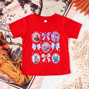 May include: A red t-shirt featuring a design of cartoon characters and bows. The design includes images of girls and a blue cat, framed by decorative bows in various colors. The shirt is made of a soft material.