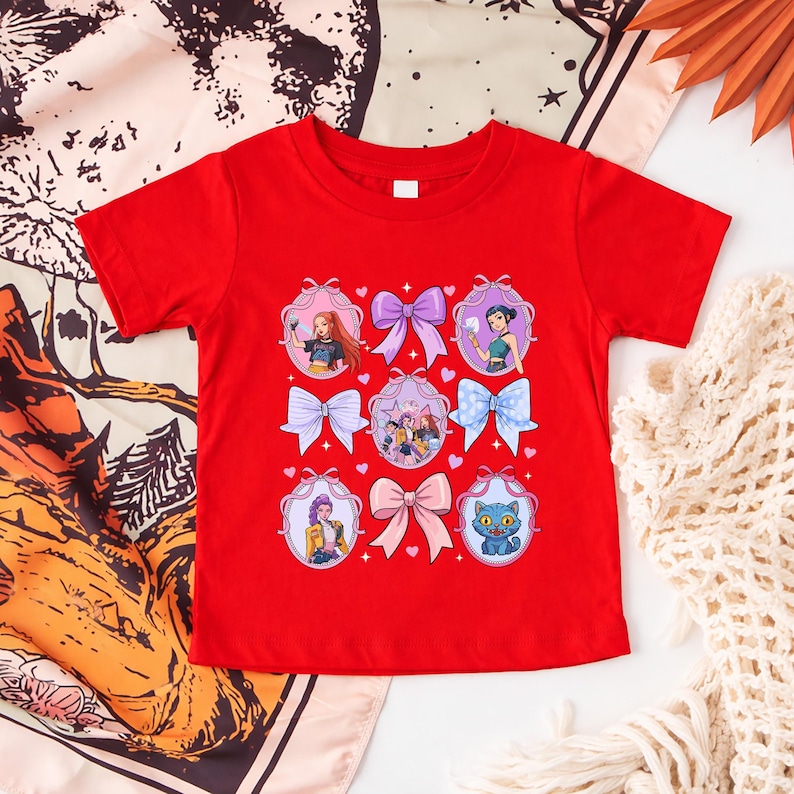 May include: A red t-shirt featuring a design of cartoon characters and bows. The design includes images of girls and a blue cat, framed by decorative bows in various colors. The shirt is made of a soft material.