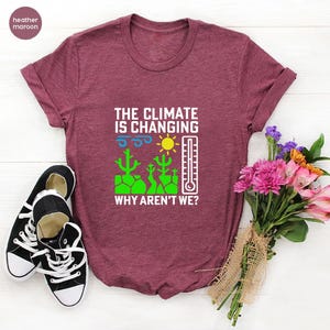 May include: Heather maroon t-shirt with the text "THE CLIMATE IS CHANGING WHY AREN'T WE?" and a graphic of a sun, thermometer, and cacti. Black and white sneakers are on the left. A bouquet of flowers is on the right.