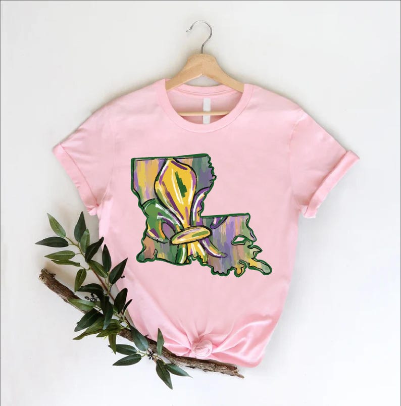 May include: A light pink t-shirt featuring a colorful illustration of the state of Louisiana with a fleur-de-lis design. The graphic incorporates shades of green, yellow, and purple. The shirt is hanging on a wooden hanger.
